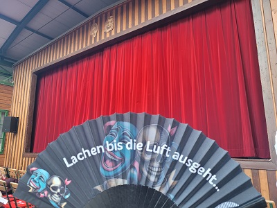 Fächer mit Aufschrift "Lachen bis die Luft ausgeht ..." vor rotem Samtvorhang der Tschauner Bühne