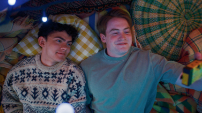 Ein Junge mit dunklen, lockigen Haaren (Charlie) neben seinem Freund mit blonden, glatten Haaren (Nick). Die beiden liegen im Bett und machen ein Selfie.