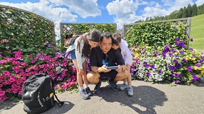 Papa mit Töchtern bei der Talstation der Gemeindealpe vor bunten Blumen mit Rucksack. Alle blicken auf ein Smartphone, wo ein Escape Trail Video läuft