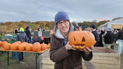Mann mit Kürbis in der Hand, der zu einem gruseligen Gesicht geschnitzt ist im Hintergrund Menschen und Kürbisse beim Kürbisfest am Himmel