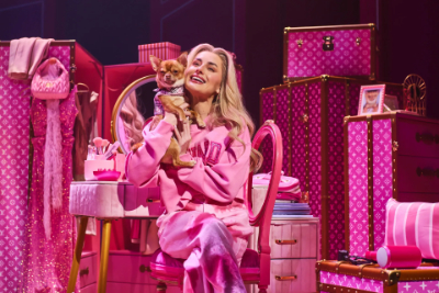West End Highlight #1: Eine weiße blonde Frau (Elle Woods) hält in einem rein pinken Raum ihren Hund in der Hand in Natürlich blond