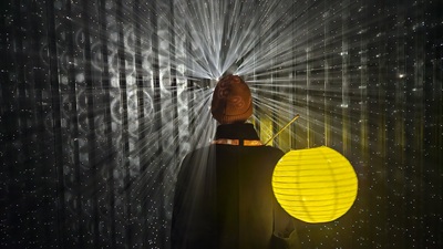 Lichtstrahlen kommen aus einer Wand, davor steht eine Frau mit gelber Laterne