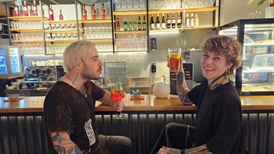 Ein Mann trinkt mit Strohhalm ein Spritzgetränk, eine Frau lacht mit Bier in der Hand. Sie sitzen an der Bar bei Golfrausch im Auhof Center.