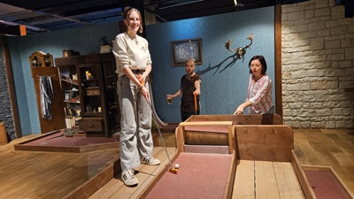Drei Personen spielen Minigolf - im Hintergrund ein Wohnzimmerschrank als Bahn.